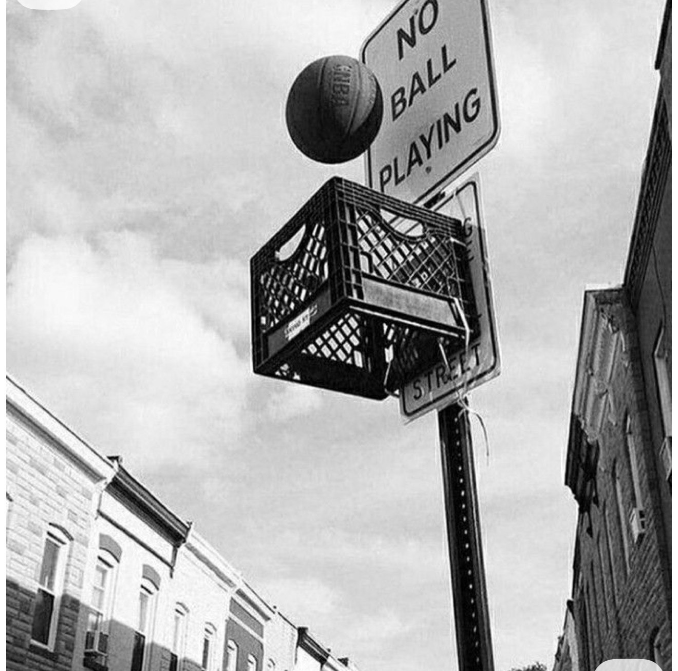 Black and white street hoop: milk crate bolted to a 'No Ball Playing' sign, urban rowhouse backdrop