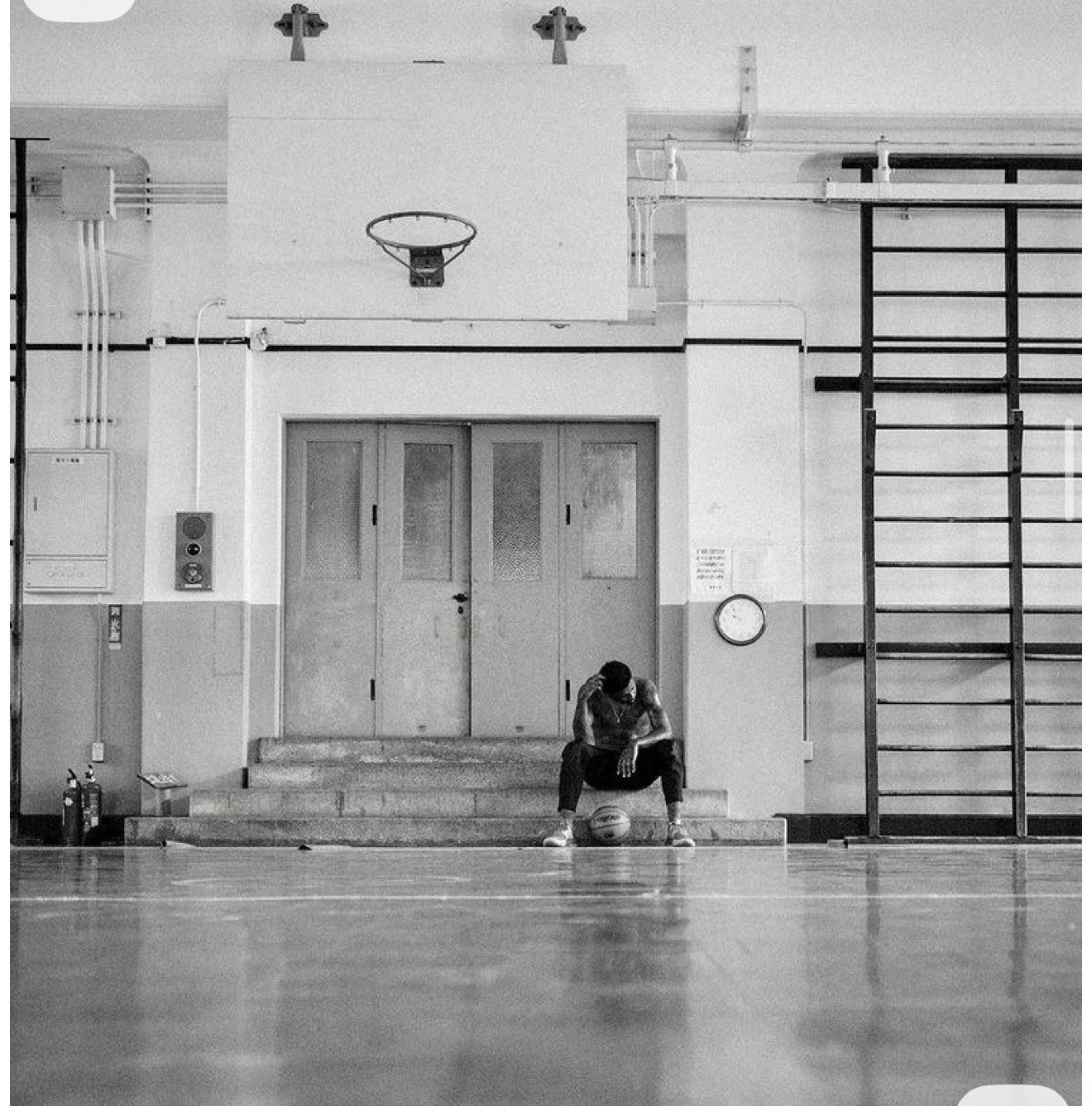 Single athlete sitting on gym steps under an austere wall-mounted hoop, documentary black and white