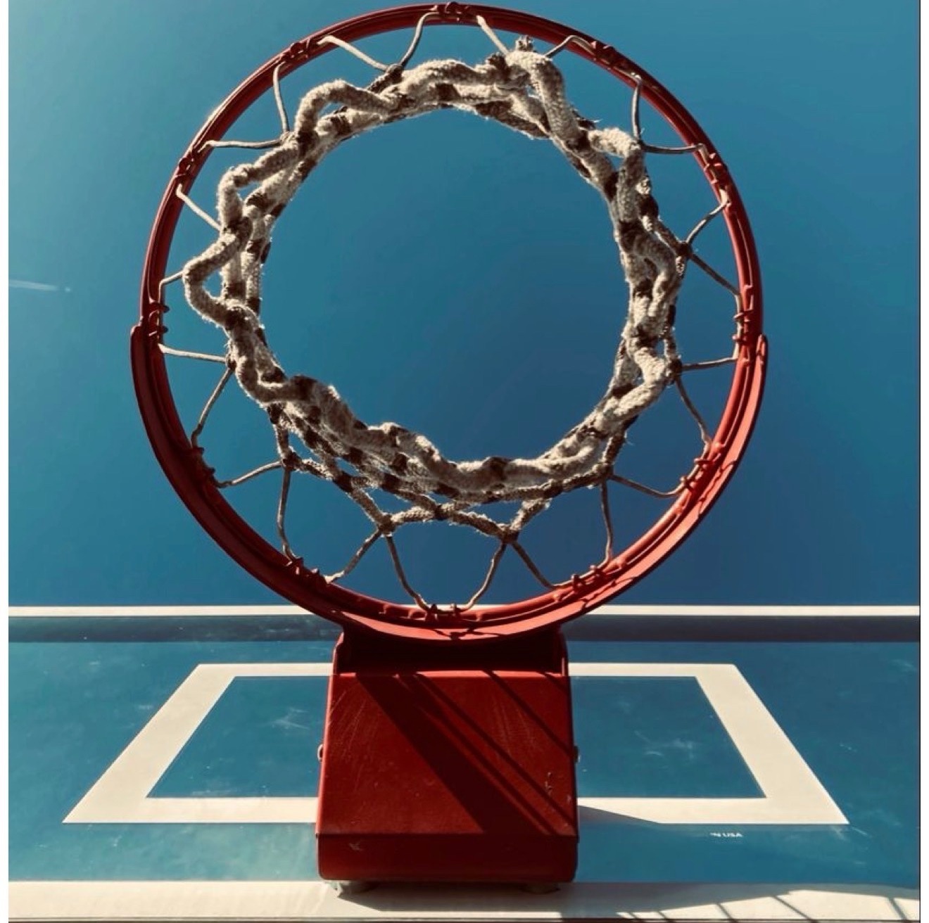 Red outdoor basketball rim with a worn knotted white net against a teal backboard and blue sky